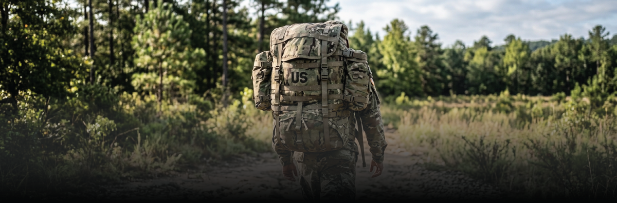 A person wearing military-style camouflage gear and a large backpack walks along a dirt path through a wooded outdoor setting.