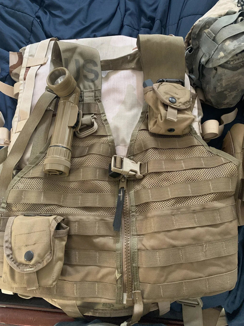 A tan tactical vest with MOLLE webbing, front zipper, and multiple utility pouches, laid flat on a dark blue fabric surface with other gear partially visible around it.