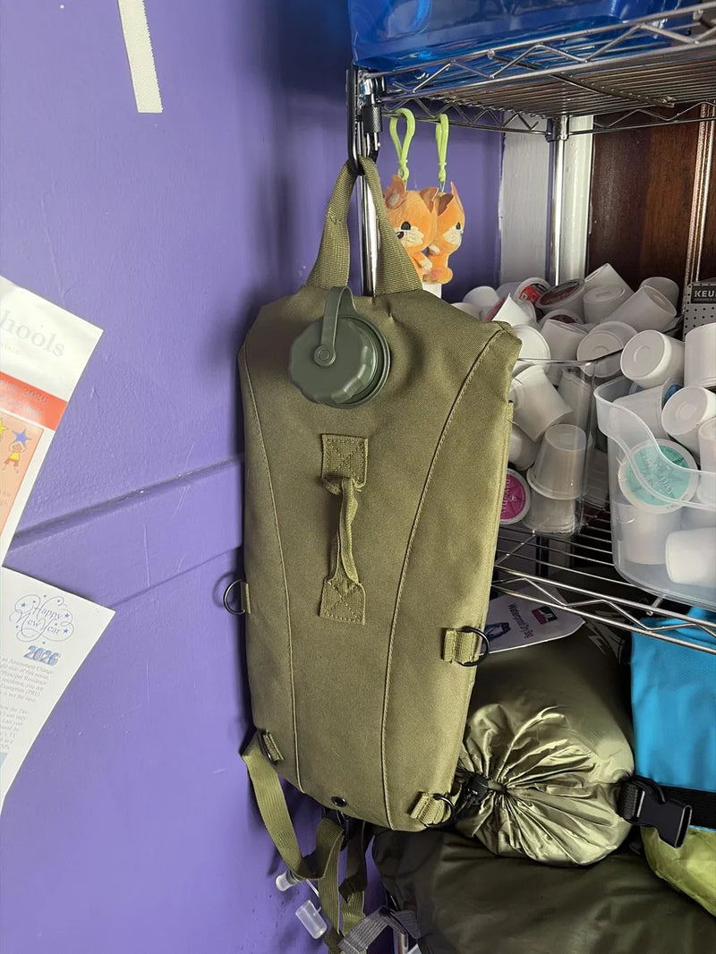 An olive green tactical backpack with a water bottle attached, hanging on a metal shelf next to a purple wall.