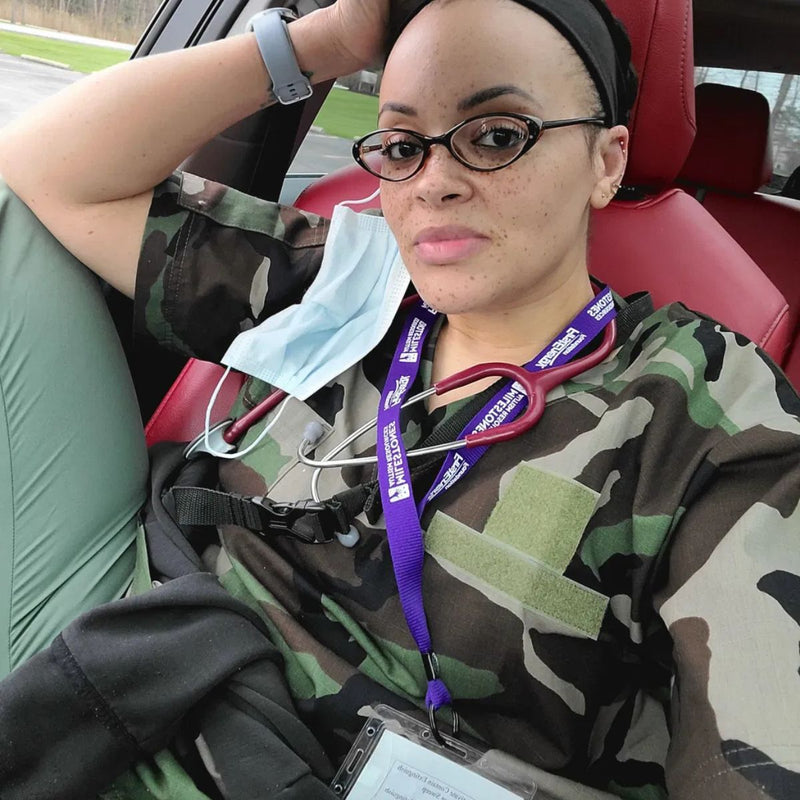 A woman wearing a camouflage scrub top and a purple medical ID lanyard sits inside a red car. She is wearing black-rimmed glasses, a black headband, and has a light blue face mask hanging from her shoulder.