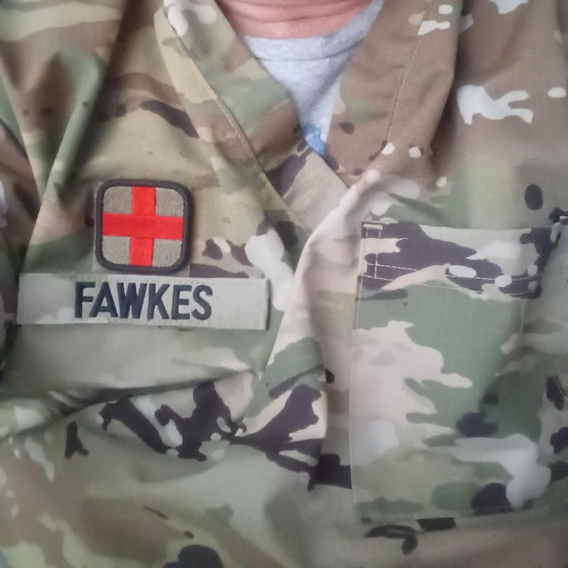 Close-up of a camouflage military-style uniform with a name tape reading FAWKES and a small patch featuring a red cross on a gray background.