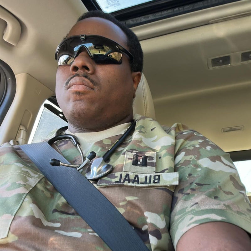 A man wearing a camouflage military-style shirt and black sunglasses, sitting inside a vehicle with a seatbelt across his chest.