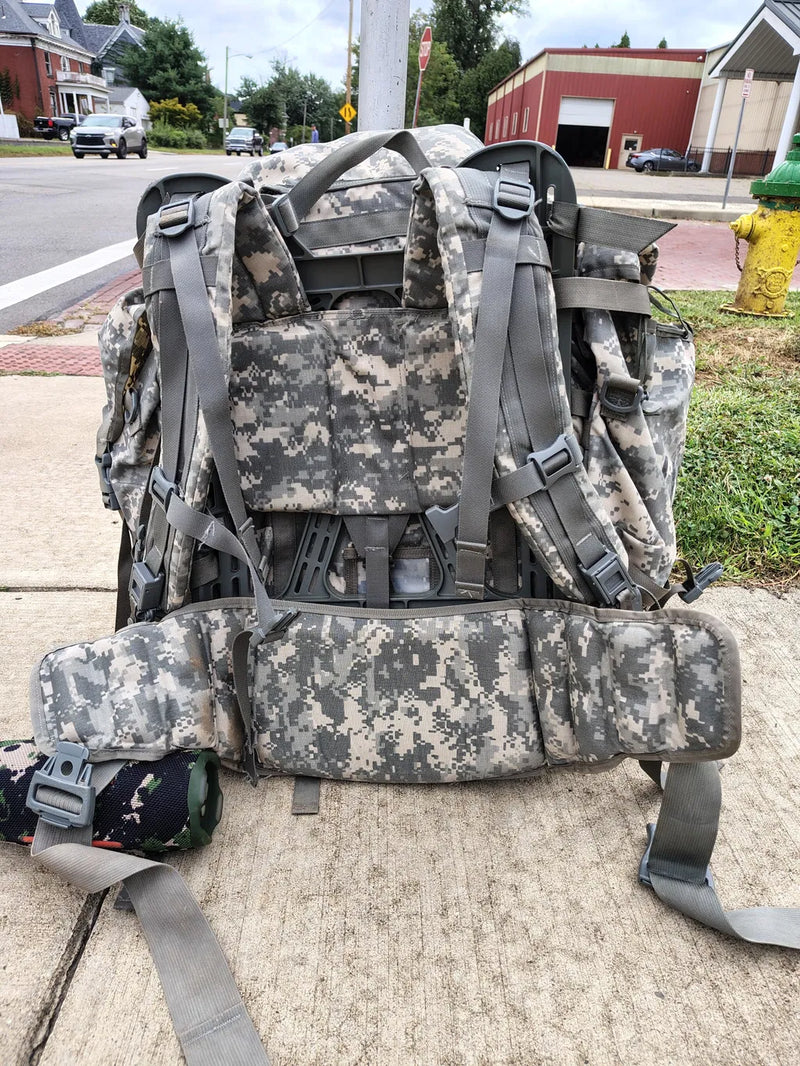 A large military-style backpack in digital camouflage pattern, photographed outdoors on a sidewalk with a street, buildings, and hydrant visible in the background.