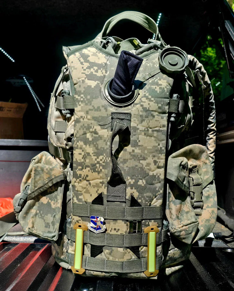 A tactical backpack in digital camouflage pattern, shown in a vehicle bed at night with its front flap open, revealing a black pouch, straps, and two bright yellow-green handles.