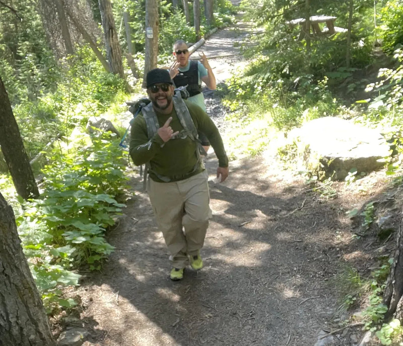 A man hiking on a sun-dappled forest trail wearing an olive green long-sleeve shirt, khaki pants, and a backpack, with another hiker behind him in a wooded outdoor setting.